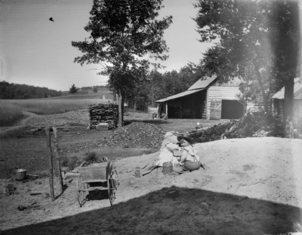 View from the shade of a building towards a boy and girl sitting on a pile of dirt playing. There is a small buggy on the left. In the background are a number of wood piles. On the left is a field with trees on a hill in the background, and on the right, farm buildings among trees.