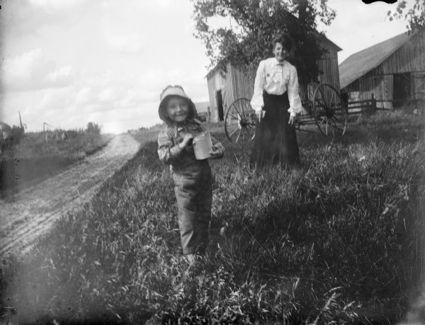 A girl wearing a bonnet is holding a can in her arms while standing in the grass along a dirt road. Just behind her is a woman, wearing a white blouse and long black skirt, standing and looking at the girl. Behind them is a horse-drawn agricultural implement parked in the grass, and beyond are farm buildings.