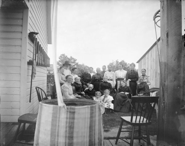 Large group of women and young girls, and one man, posing between outdoors between two buildings. In the foreground is a barrel and chairs.