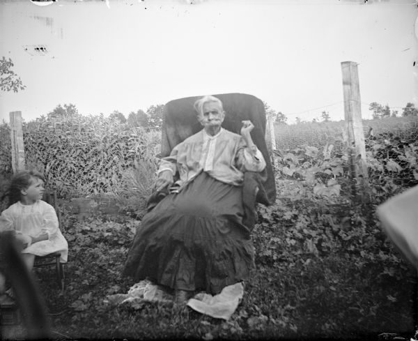Portrait of an elderly woman, and a young girl on her left, each sitting in a chair in front of a wire fence. Behind the fence is a cornfield.