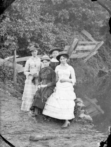 Four women posing standing together near water. Behind them is a fence angled steeply on a bank covered with foliage.