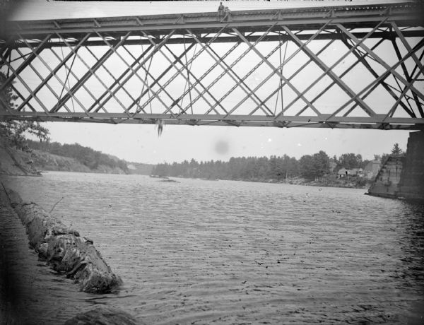 View across river towards a swimmer, caught in mid-air, jumping from the railroad bridge. Two people are on top of the bridge looking down.	In the background on the right riverbank are buildings.
