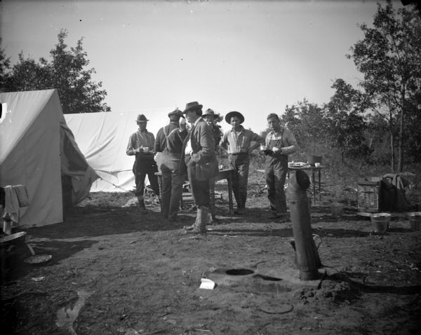 Six men posing standing around two tables next to two tents. In the foreground is a stovepipe coming out of the ground.