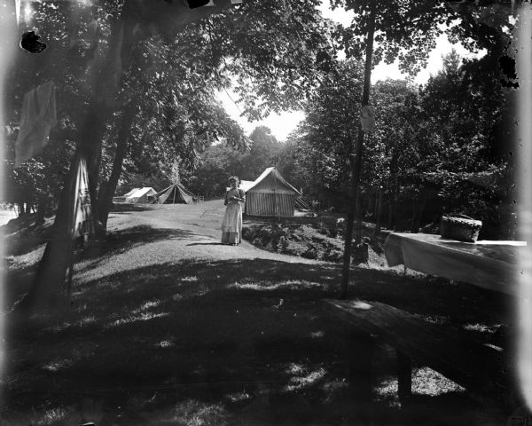 View across lawn towards a woman posing standing in front of several tents. Next to her on the right is a ravine. In the right foreground is a picnic table. On the far left in the background is a river or lake.	