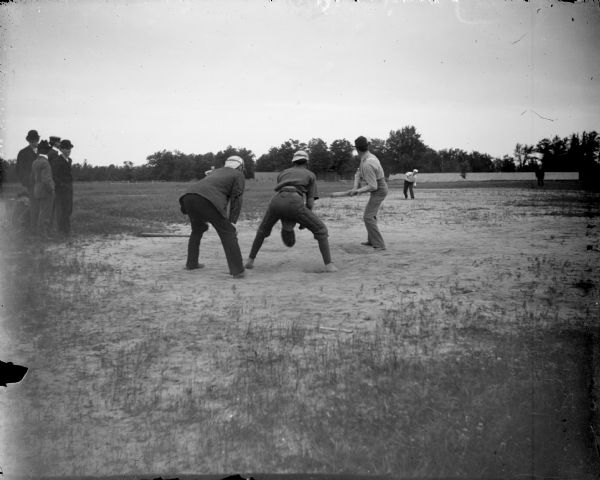 Pitcher, batter, catcher, outfielder and umpire at a baseball game, with four spectators on the left.	