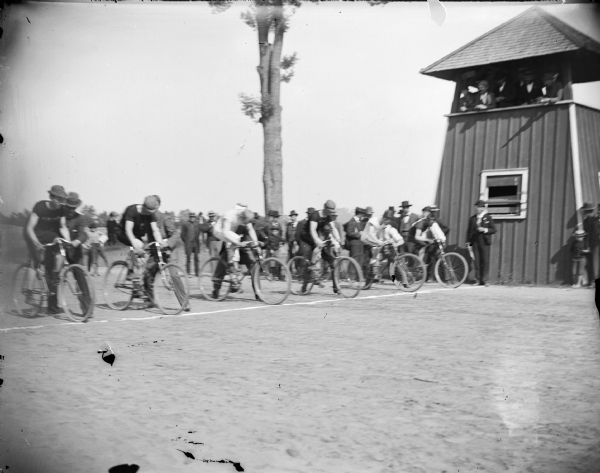 Cyclists at the starting line of a race near the judges' stand. Each cyclist has a man standing next to him holding the bike upright for the start of the race. A crowd of people are in the background.