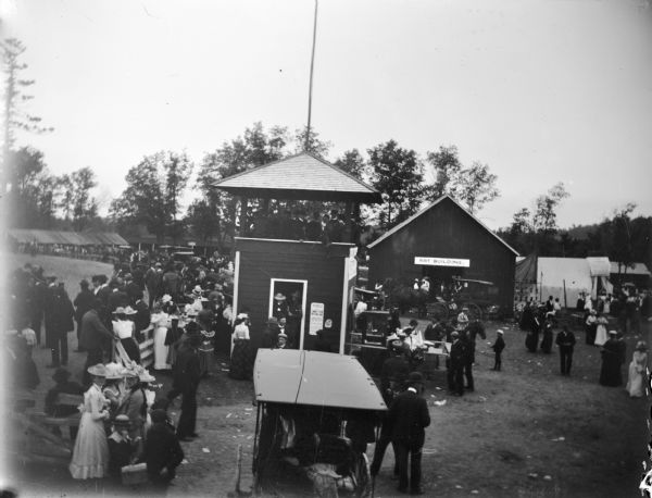 Elevated view of the scene at the country fair. In the foreground a crowd is gathered near a pavilion near the racetrack. In the background on the right is the "Art Building." There is a horse-drawn carriages and in the foreground. Other fair buildings are in the background.