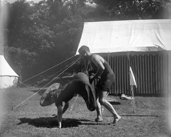 Man and woman in swimming outfits in a wheelbarrow position in front of tents.