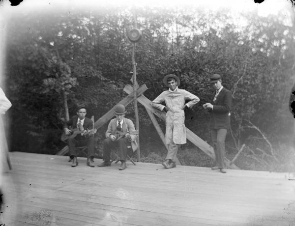 View across stage towards two men posing sitting, one playing a guitar and the other a mandolin, with two other men standing nearby. In the background are trees. A woman wearing a dress is partially seen on the far left.