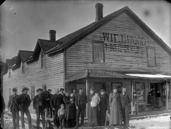 Large group of men, women, children and dogs posing standing in front of the Wildwood Hotel. Snow is on the ground.