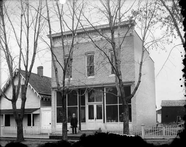View from street towards a man posing standing and holding his hat at the entrance of a two-story brick building with a storefront. There is a wood frame house on the left. A white picket fence is in front of the house and storefront and side yard on the right. In the background on the right is a barn.