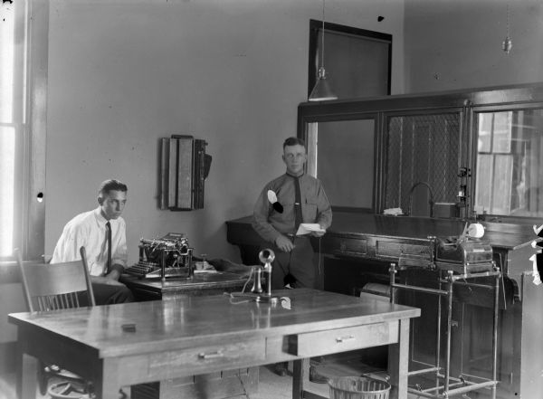 Man posing sitting and a man posing standing in a office behind a counter with a wire window for customers. Equipment in the office includes an adding machine, telephone and typewriter.