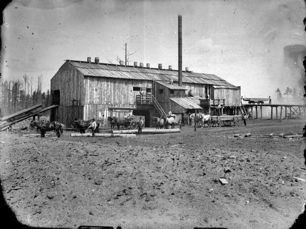 View across open ground towards men posing sitting and standing in horse-drawn wagons in front of a sawmill.	