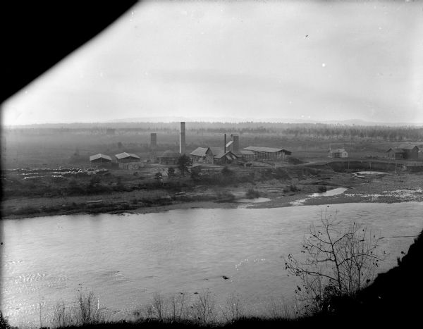 Elevated view from opposite shoreline of Halcyon Brick Works on the shore of a river. Trees, fields and hills are in the far background.