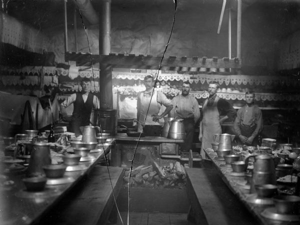 Five men are standing in a row behind a stove and long tables. Interior of what may be a lumber camp cook shack.