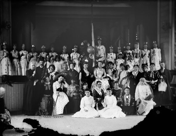 Large group portrait of men, women and children dressed in costume posing in front of, and on a stage. Probably a theatrical cast.