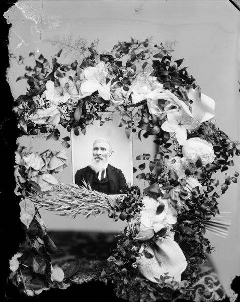 Studio portrait of a flower arrangement surrounding a studio portrait of a man with a beard and wearing a suit.