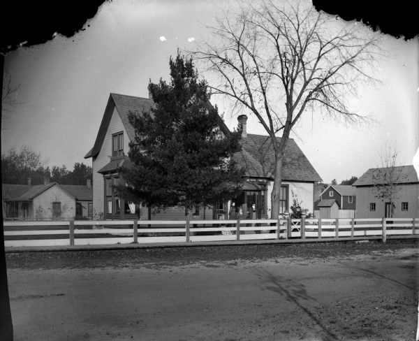 View across unpaved road and fence towards five women. Four of them are posing standing, and a woman is sitting in a yard in front of a two-story frame house in town. Other houses and outbuildings are in the background on the left and right.