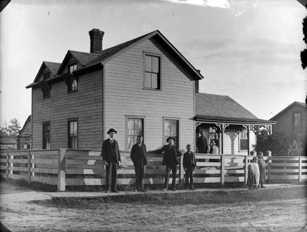 View across unpaved road towards four boys and three girls posing standing in front of a fence in front of a two-story house. A man, and a woman holding a baby, are posing standing on the front porch.