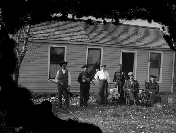 Group portrait of men, women and children posing in front of a single-story frame house. The man on the left is holding a shotgun, the man next to him is holding a violin, and two women in the center are standing near an infant in a baby carriage. An older man with a pipe, and woman holding a puppy, are sitting in chairs on the lawn. A young boy sitting is between them on a stool.