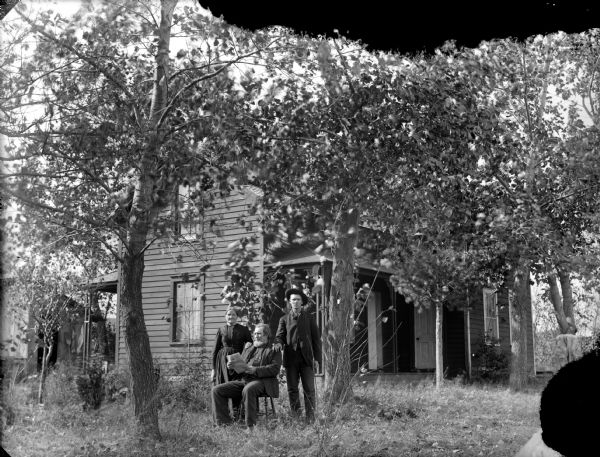 Man posing sitting and reading a newspaper between a man standing man on the right, and a woman on the left. Behind them is a frame house among trees.