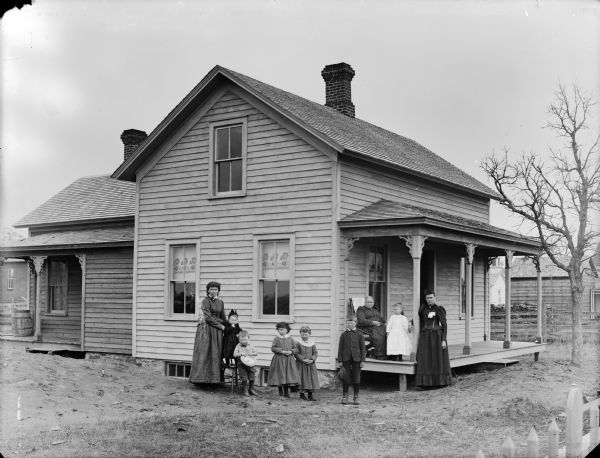 Two women, two boys, two girls, and a small girl are posing standing at the corner of a frame house. A woman is standing next to a column of the porch, and on the porch is a woman posing sitting, and a small girl standing. In the background are other buildings.