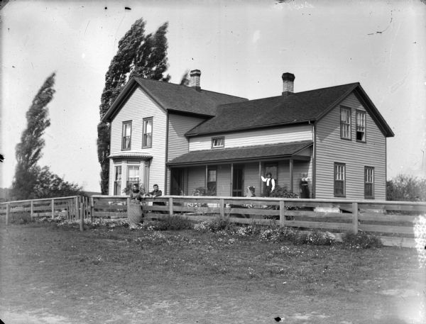 Two women are posing standing on either side of a wooden fence in front of a two-story frame house. A man and woman are posing standing on the porch.
