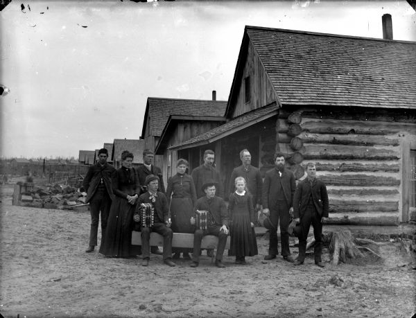 Two men are posing sitting on a bench holding accordions. and six men, two women, and a girl are posing standing around the two men. In the background is a log building.