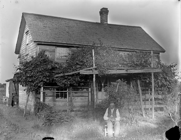 Elderly man posing sitting in front of an arbor and a two-story frame house.