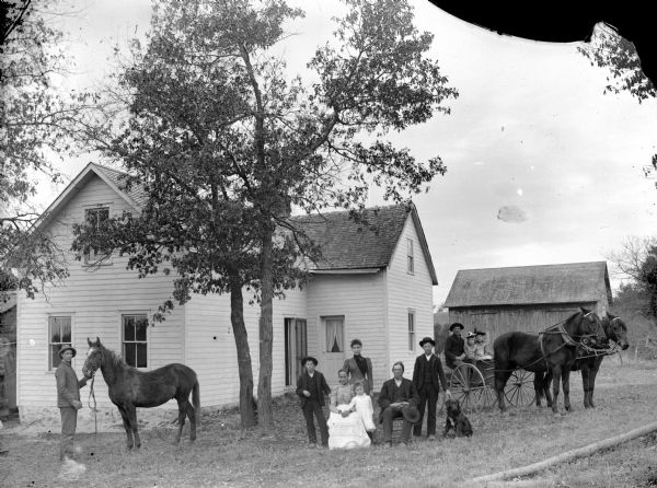 A man is displaying a horse on the left, a  man and woman are posing in the center, sitting in chairs. The man and woman are surrounded by two boys, a woman, girl, and dog, and two girls and a man are posing sitting in a buggy pulled by a team of two horses in front of a two-story frame house.