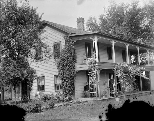 View across lawn towards a man posing standing and a woman sitting on the porch of a two-story frame house with a balcony above the porch.