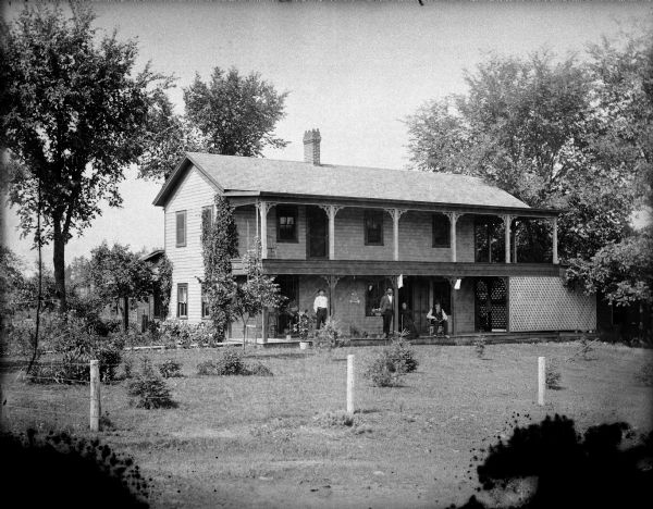 View across lawn towards a man and woman posing sitting, and two men standing on the porch of a two-story frame house with a balcony above the porch.