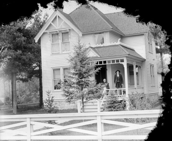 View over fence towards a man posing standing and a woman sitting on the porch of a two-story frame house. Laundry is hanging on a line in the yard on the left.