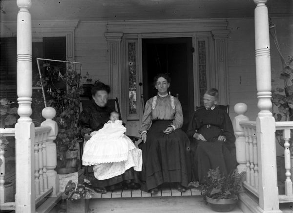 Three women are posing sitting on a porch in rocking chairs in front of the door of a house. The woman on the left is holding an infant wearing a baptismal gown.