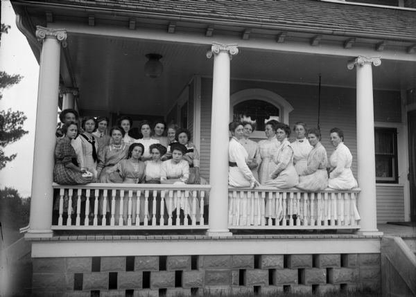 View from yard of twenty women posing standing and sitting on the porch of a house.