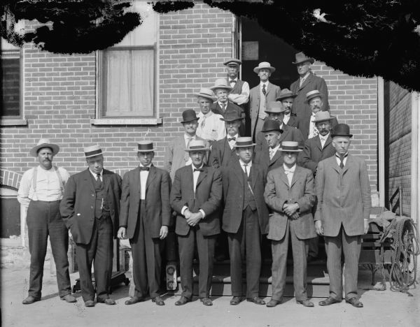 Outdoor group portrait of men dressed in suits posing standing on the sidewalk and steps in front of the brick building that houses American Express and the photography studio of Charles J. Van Schaick.