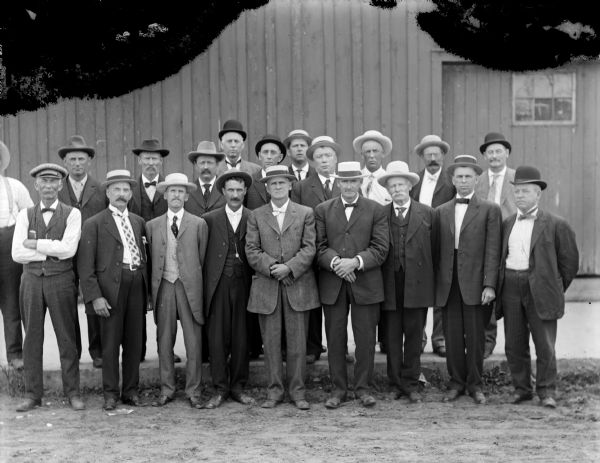 Outdoor group portrait of men dressed in suits posing standing in the street and on the curb in front of a wooden building.