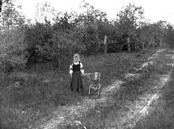 Girl pos700011270039 standing next to a stump next to a small baby stroller with a doll in it, on the side of a dirt road or path. Behind the girl are bushes along a wire and wood fence line.