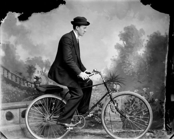 Studio portrait in front of a painted backdrop of right side profile view of a man posing sitting on a bicycle.