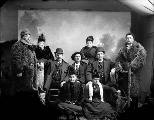 Studio group portrait of women and men posing sitting and standing. They are all wearing hats and coats. Two of the men wear thick, fur coats. Against the back wall is a painted backdrop, and part of a skylight is on the upper right.