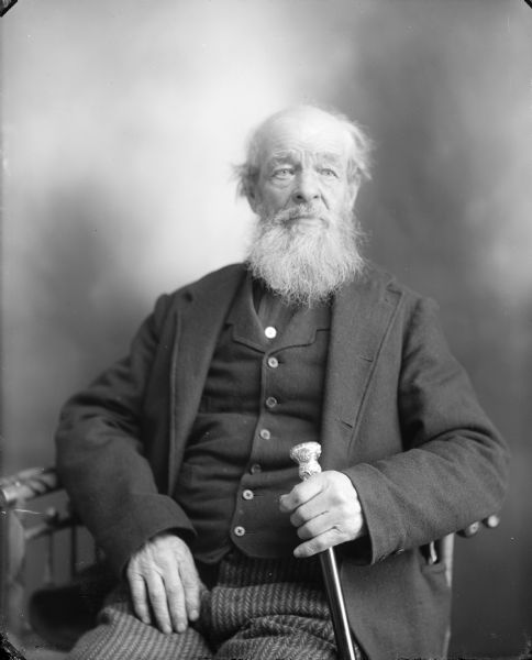 Studio portrait of an elderly man with a beard posing sitting and holding a cane.