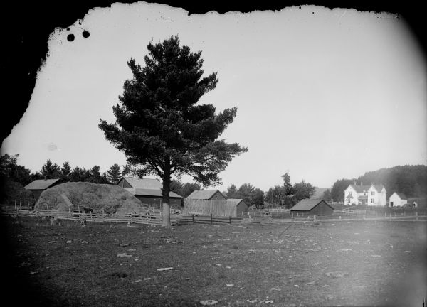 View across field towards farm buildings. In the foreground are tree stumps, a large tree, and fences, a haystack, and wagons. There is a two-story frame house in the background on the right. Behind the farm buildings are trees.