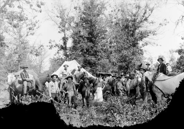 Large group of men posing standing among other men on horseback in front of tents in the woods. The horses are wearing yokes. There appears to be a wood structure with chimneys on the roof in the background on the right. One of the men in front is holding a long stick. Another man in the center is wearing a long apron.
