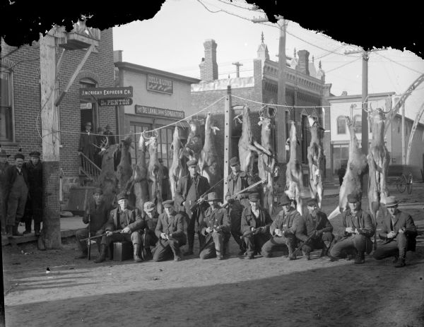 Group portrait of a dozen men posing sitting and kneeling while holding rifles in front of thirteen deer strung up across First Street. They are in front of the brick building that held Charles J. Van Schaick's Photography Studio, American Express, and Dr. Penton. Other men are posing on the stoop of the building, and also on the sidewalk. Another storefront on the right is for Dell & Lund, Real Estate Loans and Abstracts.
