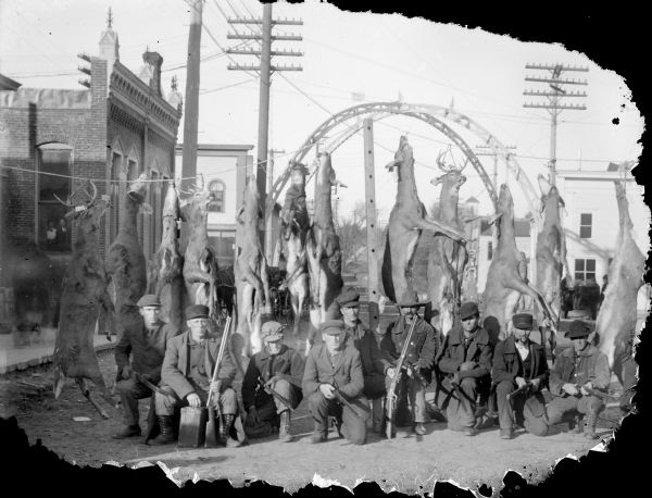 Nine men posing while holding rifles in front of twelve deer strung up across First Street in front of the brick building that held Charles J. Van Schaick's Photography Studio. In the background is a metal arch across the intersection.