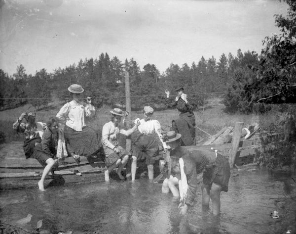 Eight women posed wading and playing in a river. Some of the women are sitting on a wood fence across the river.