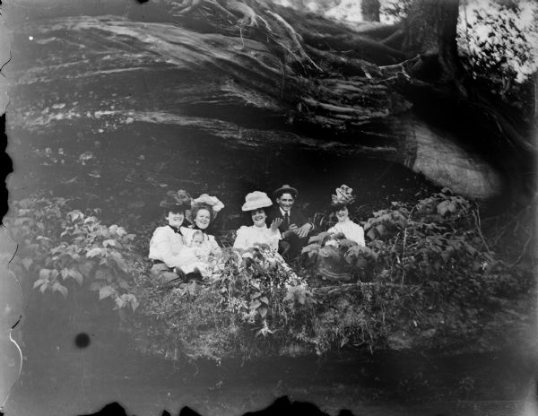 Four women, an infant, and a man are posed sitting together on a grassy ledge under a rock ledge. Above them are trees and tree roots.