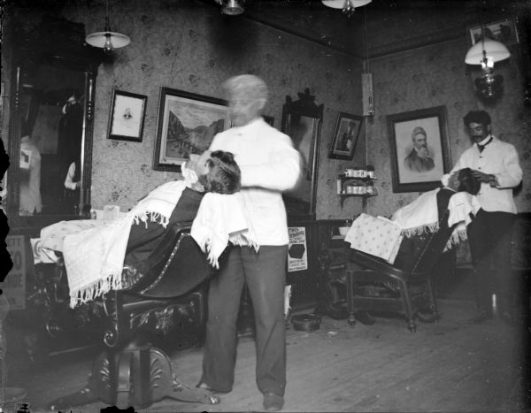 Interior of a barbershop. Two barbers are shaving two men.