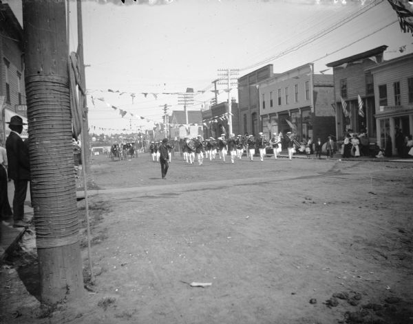 View down unpaved street towards a marching band parading up Main Street during a patriotic celebration. Pedestrians watch from the sidewalks.