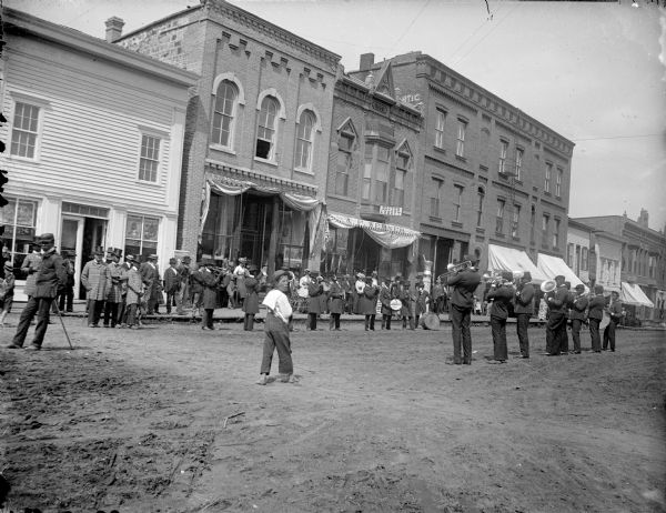 View across unpaved Main Street towards an African-American brass band standing and playing in front of the storefront for A.F. Werner. Pedestrians watch from the sidewalk.